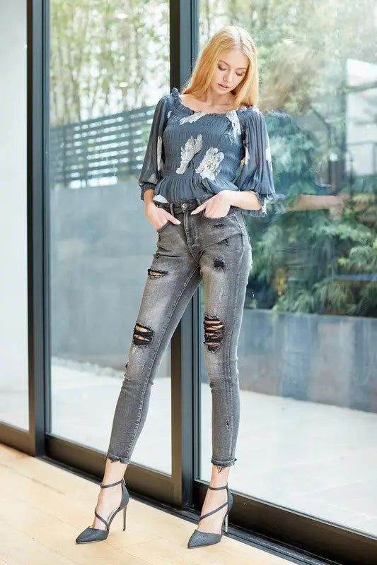 Woman modeling distressed gray mid rise skinny jeans, patterned blouse, and black stilettos