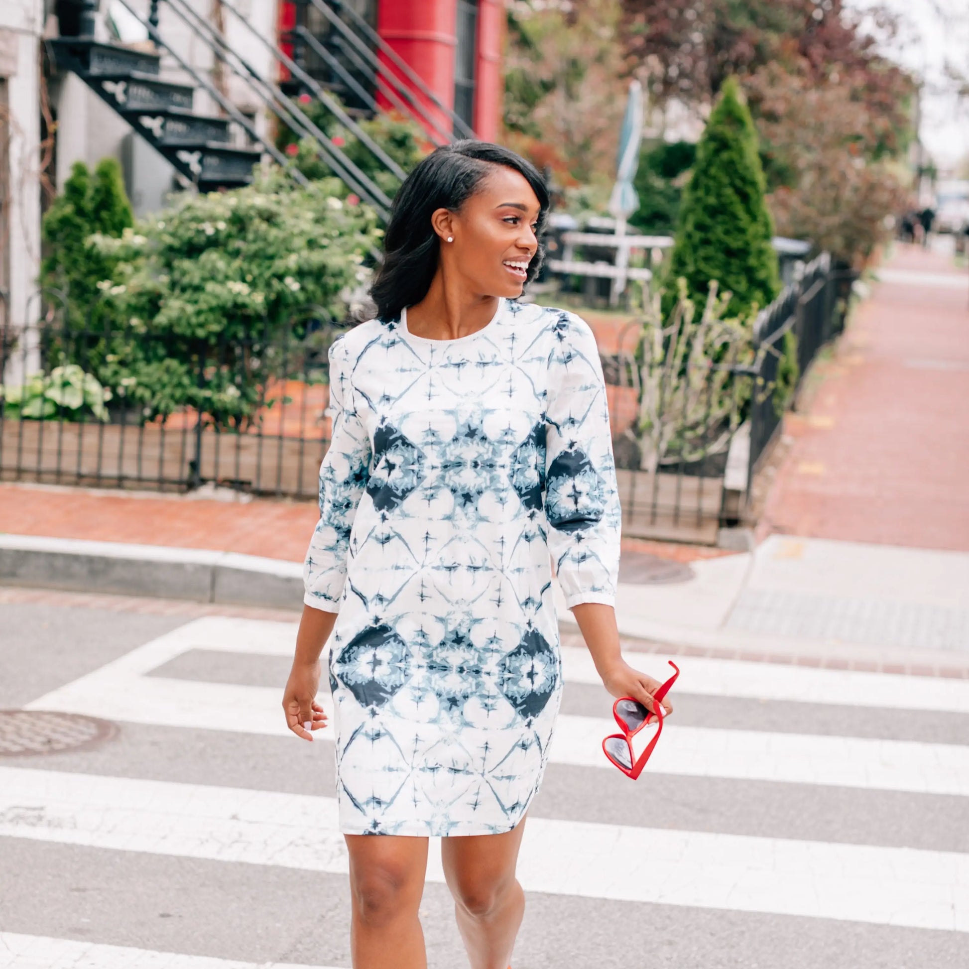 Woman in a white and blue woven slim fit dress holding red heart-shaped sunglasses