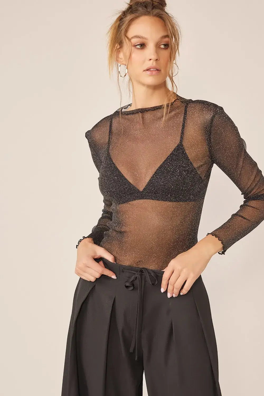 Woman wearing Idem Ditto sparkling glitter long sleeve sheer top with black bra and pleated pants
