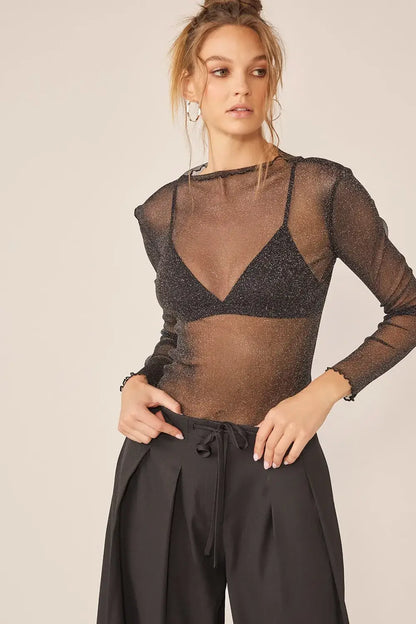 Woman wearing Idem Ditto sparkling glitter long sleeve sheer top with black bra and pleated pants