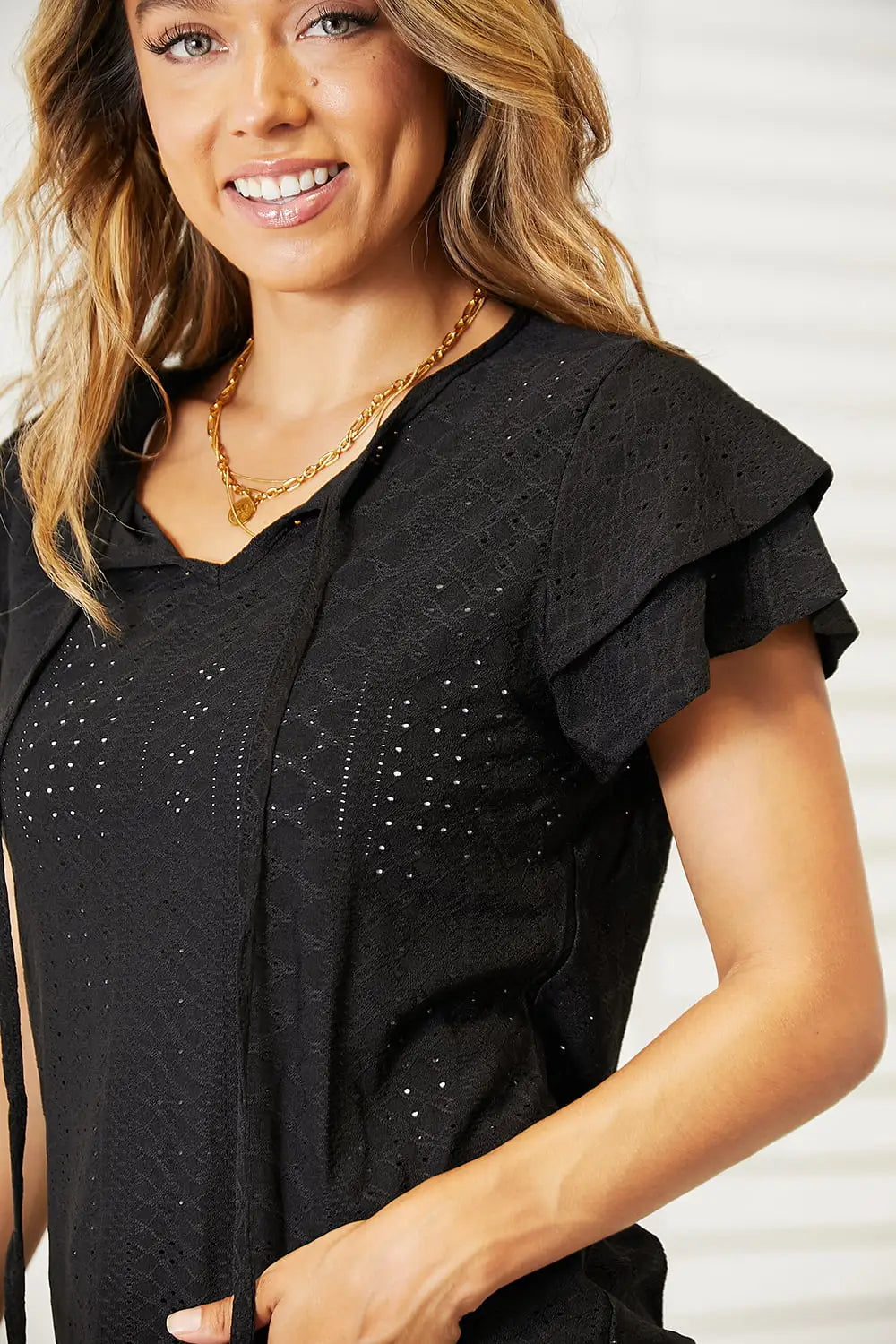 Black flutter sleeve blouse with eyelet tie-neck, gold necklace, and bracelet