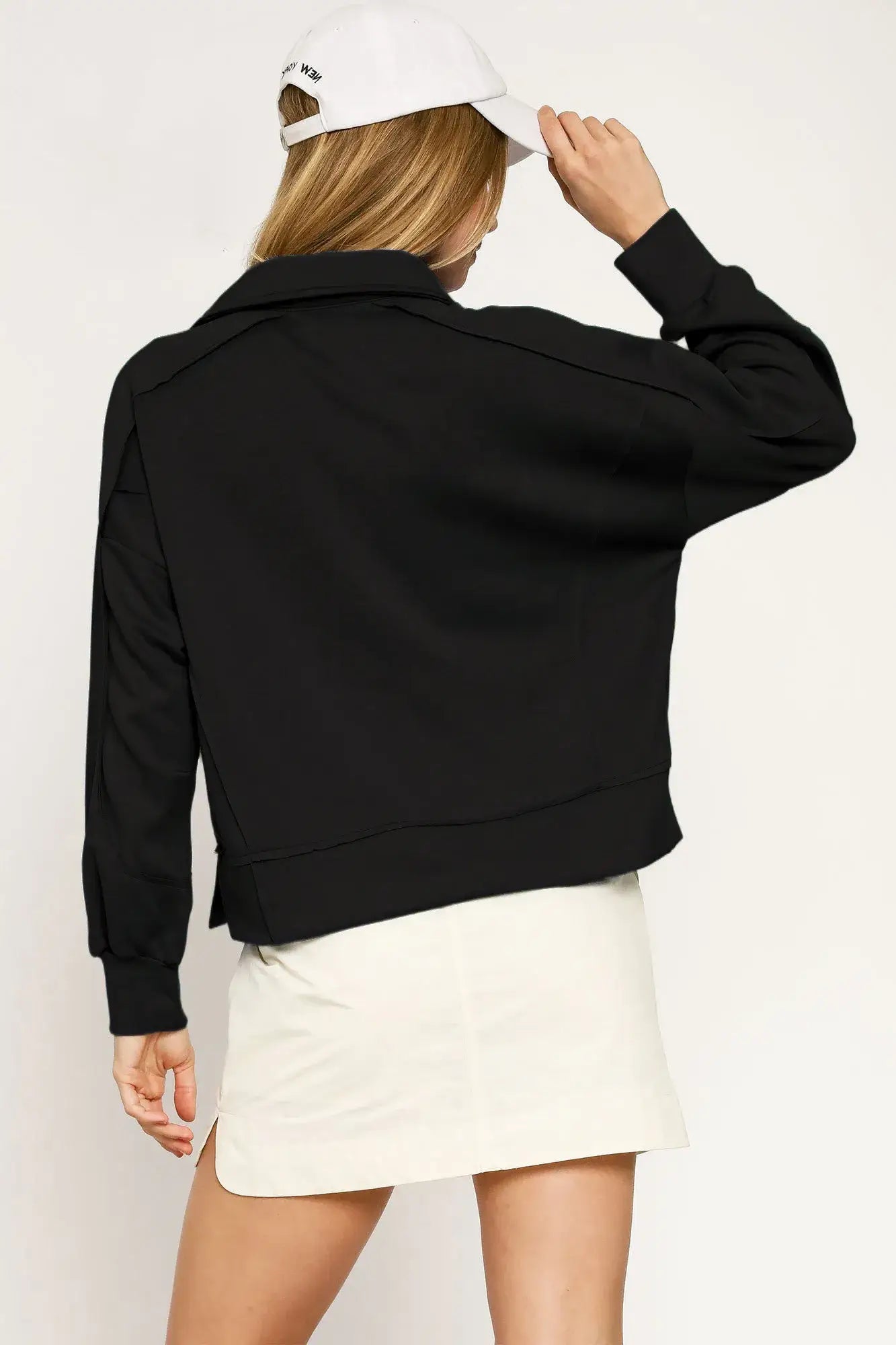 Black cropped bomber jacket with collared neck and raw hem detail, paired with a white cap and cream skirt