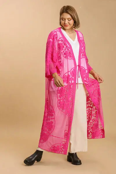Woman wearing bright pink floral lace open kimono with waist tie over white top and cream pants